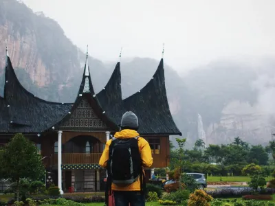 A hiker stands in front of an old asian building. Misty mountains in the background.